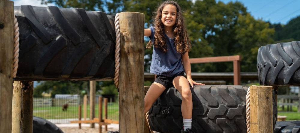 The image shows a young girl smiling brightly as she sits on top of a large stack of tires in an outdoor playground or obstacle course. The tires are supported by sturdy wooden posts and ropes, creating a fun climbing structure. The girl is wearing a blu