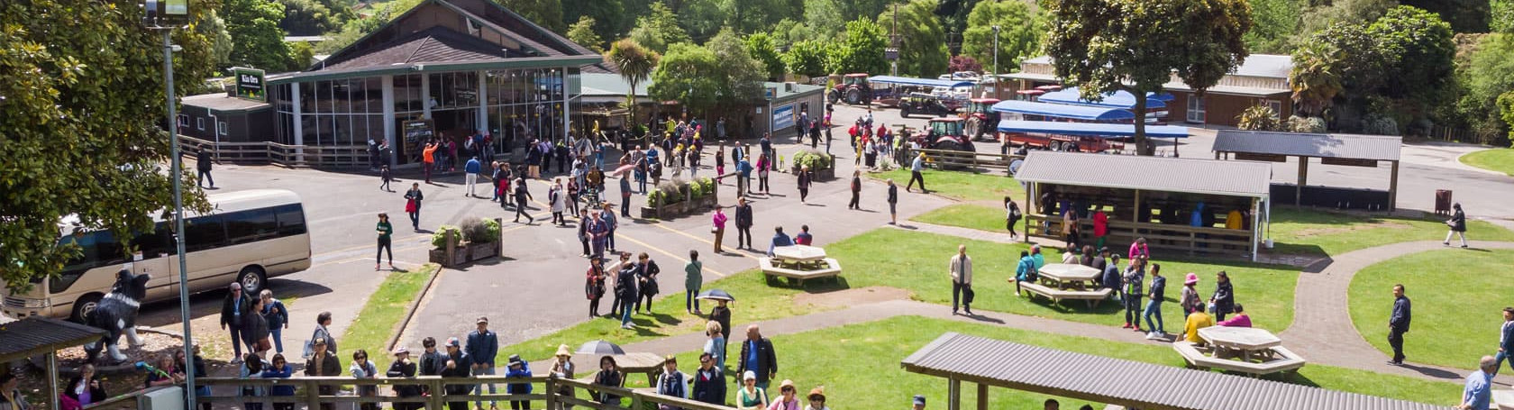 The image shows a lively outdoor scene at a farm or park, with numerous people walking, gathering, and exploring the area. In the foreground, visitors stand along a wooden fence, possibly watching an animal demonstration. The mid-ground features picnic ta